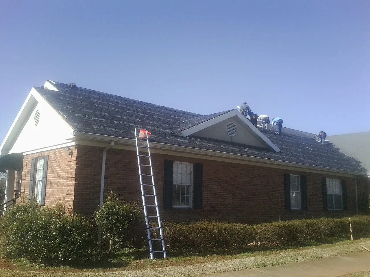 Crew tearing shingles off a brick ranch roof with a ladder set — Emergency Roof Repair work by our crew in Chichester